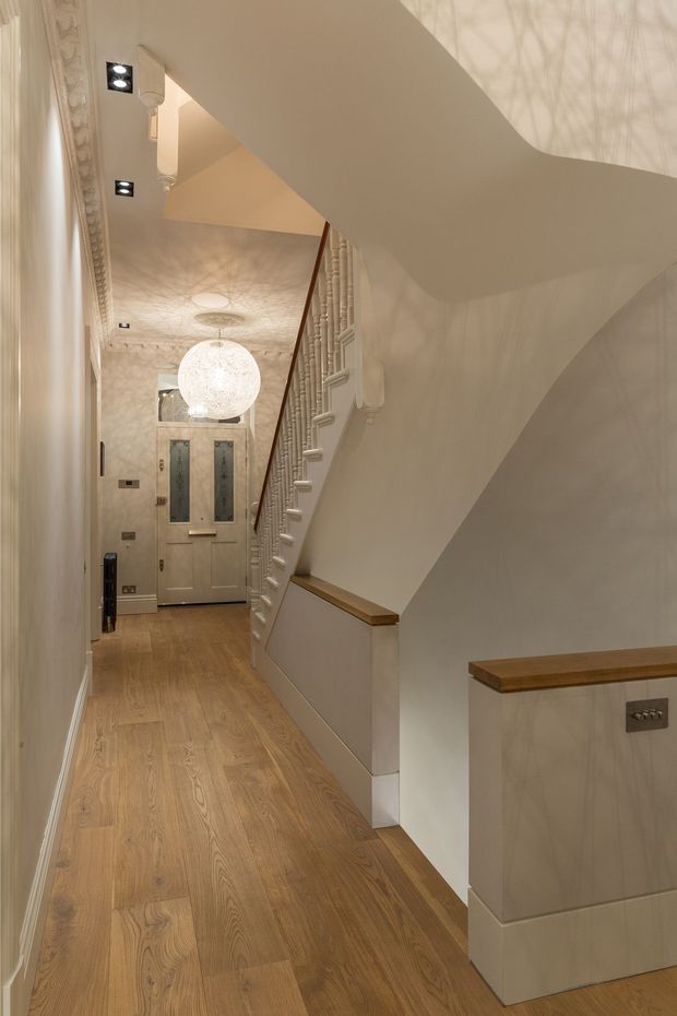 A refined residential hallway and stairway with warm timber flooring, pale painted walls, ornate decorative mouldings, a sculptural white staircase, and a classic glazed entry door at the end of the corridor. Integrated downlights and a large spherical pendant create a softly illuminated, elegant interior.