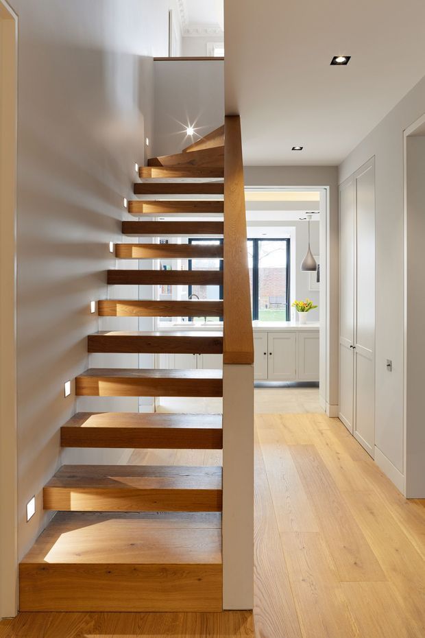 Bright contemporary interior view of a timber floating staircase with thick open-riser treads, a central timber support, and integrated wall lighting. The stairway opens onto a light timber-floored hallway with white built-in cabinetry and a glimpse of a kitchen or living area beyond. Recessed ceiling lights and restrained neutral finishes create a clean, refined atmosphere.