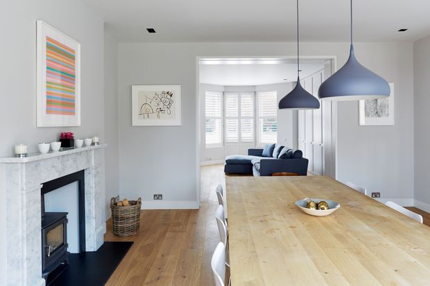 Bright open-plan dining and living interior with a long natural timber dining table, white dining chairs, pale grey walls, a marble fireplace surround with wood-burning stove, and two large blue pendant lights. A comfortable living area with a navy sofa is visible through the wide opening, with a bright bay-windowed space beyond. The room has a clean, refined contemporary-traditional feel.