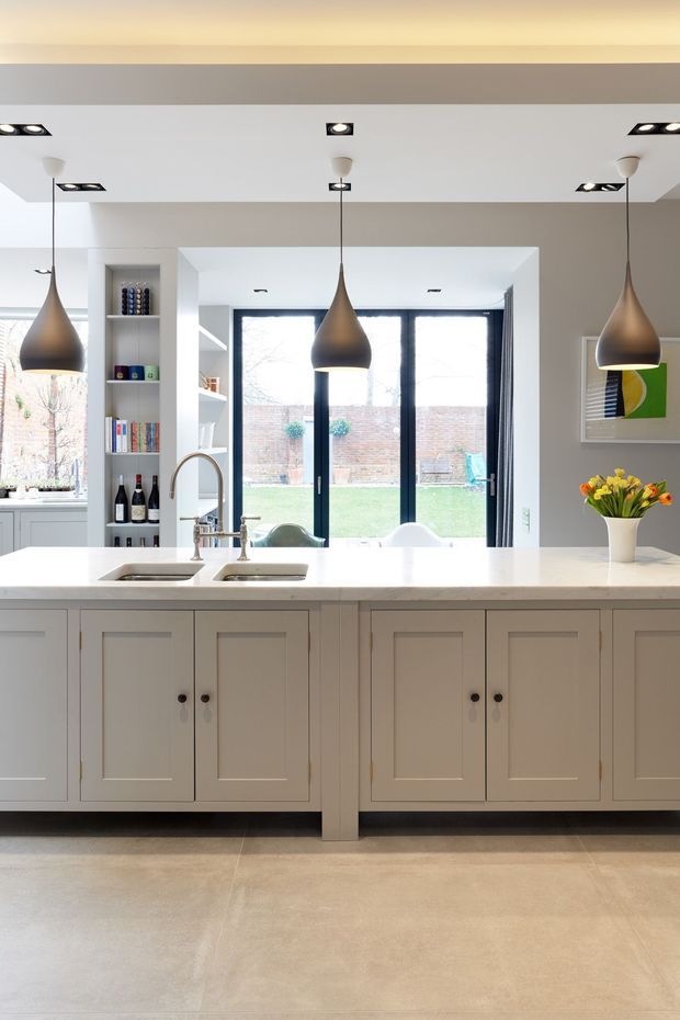 Bright, refined kitchen viewed from the living side, centred on a large painted panelled island with a pale stone benchtop, double undermount sink and brass-toned tapware. Three bronze pendant lights hang above the island, with white cabinetry, open shelving, integrated appliances, and full-height black-framed glazing opening to a garden courtyard. Pale large-format flooring and soft grey walls create a clean, contemporary-traditional look.