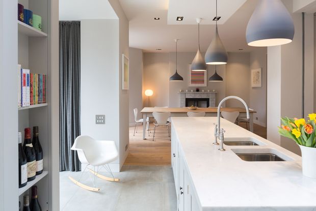Bright open-plan kitchen and dining area viewed from the kitchen island. The space features white cabinetry and benchtops, an integrated double sink with curved tap, timber flooring in the dining zone, pendant lighting, a built-in fireplace, open shelving with books and wine storage, and a white sculptural rocking chair. The palette is light and contemporary with grey curtains and warm timber accents.