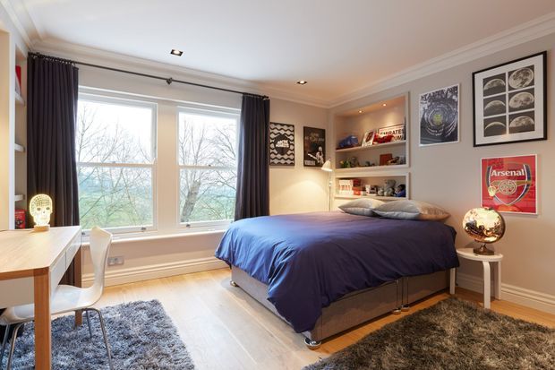A bright, well-appointed bedroom with a double bed dressed in deep blue bedding, built-in open shelving, framed wall art, and a small side table with a globe. Large windows with dark curtains overlook trees, while a white desk and chair sit in the corner. Light timber flooring, layered rugs, recessed lighting, and decorative ceiling moulding create a comfortable contemporary interior.