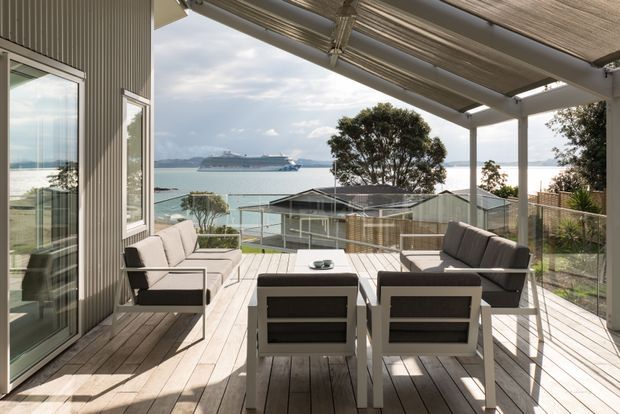 A bright elevated outdoor terrace overlooking a bay, furnished with a contemporary outdoor dining table and chairs alongside a cushioned lounge setting. The terrace has timber decking, a translucent slatted roof canopy, slim structural posts, and a vertically ribbed exterior wall with large glazed doors and windows. A cruise ship and waterfront landscape are visible beyond the glass and balustrade.