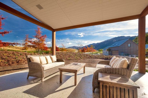 Covered outdoor terrace with wicker lounge seating and a low timber coffee table, framed by exposed timber posts and beams. The open-sided space overlooks landscaped grounds, a neighboring building, autumn foliage, and mountainous terrain under a bright sky.