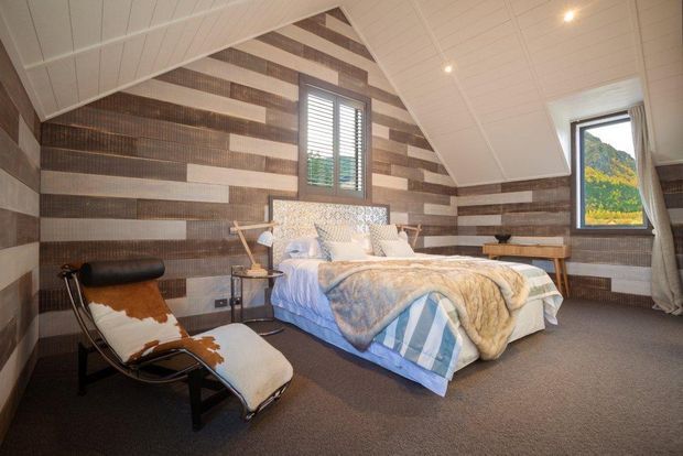 A cozy attic-style bedroom with a pitched ceiling, exposed triangular roof form, horizontal striped timber wall panelling, carpeted floor, and a central upholstered bed. The room includes a leather-and-cowhide lounge chair, bedside tables, recessed ceiling lights, and windows with views of a green mountainous landscape.