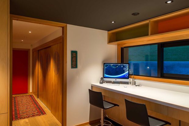 A warm, compact home office or study with a long built-in white desk beneath a wide window, two black task chairs, and a computer workstation. Honey-toned timber cabinetry and louvred storage doors line the adjoining hallway, while recessed ceiling lights and concealed shelf lighting create a moody, refined atmosphere. A patterned runner and red accent wall are visible in the adjacent corridor.
