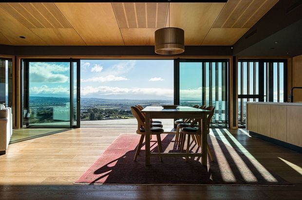 A bright, contemporary open-plan dining area with a timber dining table and chairs positioned before expansive floor-to-ceiling glazing. The room features warm timber flooring and ceiling panels, dark framing and cabinetry, a cylindrical pendant light, and strong sunlight casting linear shadows across a muted rug. A wide elevated landscape view is visible through the glass.