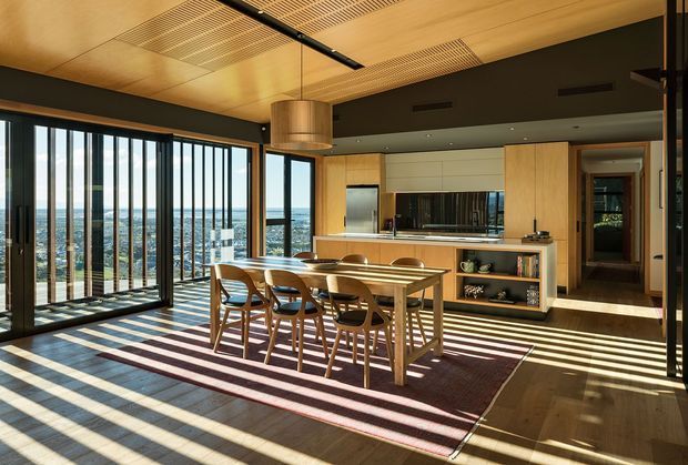 A warm, contemporary open-plan dining and kitchen area with a large timber dining table and six chairs positioned beside full-height glazed doors. The room has extensive ocean-facing views, timber ceilings with integrated linear grilles, dark walls and cabinetry accents, a light timber kitchen with island bench, integrated shelving, and strong striped sunlight across the timber floor and rug.