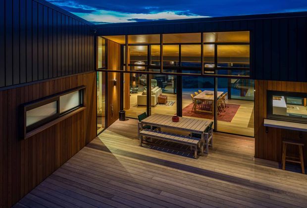 Twilight view of a modern timber-clad home centred on a large glazed corner opening. A broad timber deck forms an outdoor entertaining area with built-in bench seating, while the warmly lit interior reveals a dining area and living room through the floor-to-ceiling glazing. Dark vertical cladding, strong horizontal rooflines and extensive black-framed glazing create a contemporary architectural composition.