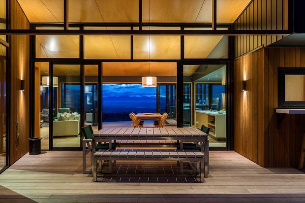 A warmly lit timber-clad outdoor entertaining deck viewed toward a glazed modern home. A large dark timber dining table with benches occupies the foreground, while floor-to-ceiling black-framed glazing reveals a dining area, living room, kitchen, and expansive blue-toned view beyond. Vertical timber wall cladding, exposed timber ceiling planes, integrated lighting, and a partially visible upper-level balustrade create a refined contemporary architectural setting.