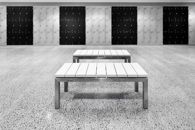 Black-and-white photograph of a spacious locker-room or changing-room interior. Rows of alternating black and white lockers line the rear wall, while two simple rectangular slatted benches sit centrally on a speckled terrazzo-like floor. The composition is symmetrical, minimal, and highly graphic.