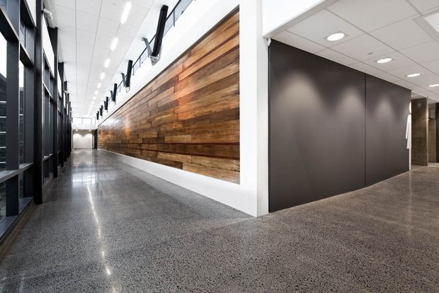 A modern commercial interior corridor with polished concrete or terrazzo-style flooring, a large feature wall clad in horizontal reclaimed-looking timber boards, and dark matte wall panels and framed glazing. The space has a bright suspended ceiling with linear and recessed lighting, creating a clean, refined office or institutional circulation area.