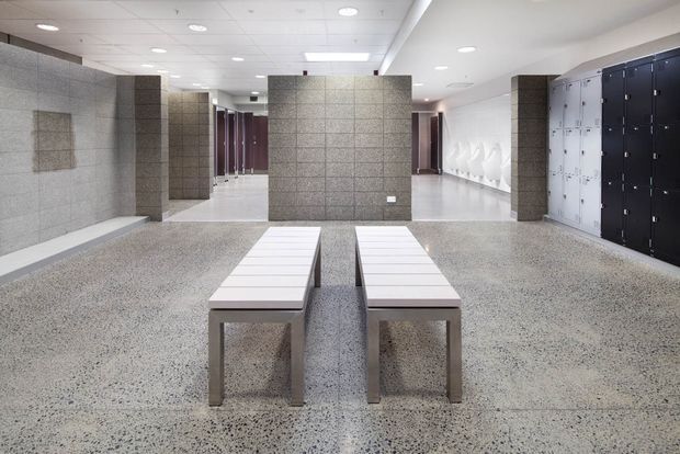 A bright, contemporary locker-room interior, likely a commercial changing facility. The space features a polished speckled terrazzo floor, grey tiled partition walls and columns, rows of black and white lockers, white wall-mounted basins or fixtures, recessed ceiling lights, and two simple white slatted benches in the foreground.