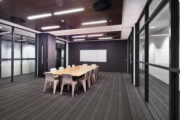 Contemporary commercial meeting room with a long light-wood conference table surrounded by white chairs. The room features dark framed glass partitions, a charcoal feature wall, grey striped carpet, and a perforated timber acoustic ceiling with linear lighting and integrated ventilation. Adjacent glazed offices or meeting spaces are visible through the partitions.