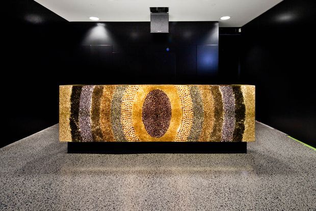 A dark, gallery-like interior featuring a large rectangular abstract artwork or decorative feature panel with concentric oval bands in gold, bronze, brown, cream, and black. The panel is mounted on a low black plinth against matte black walls, with focused ceiling spotlights and a speckled grey floor creating a dramatic exhibition atmosphere.
