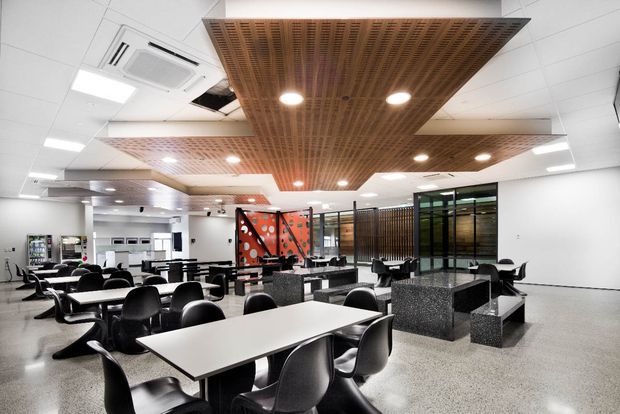 A large contemporary commercial cafeteria or staff dining area with rows of black molded chairs and white rectangular tables. The space has polished terrazzo-like flooring, white walls and ceiling panels, extensive recessed lighting, and prominent suspended warm timber slatted acoustic ceiling features. A central service or seating zone includes dark speckled stone-like counters and benches, with glazed partitions and a colourful patterned feature panel toward the rear.
