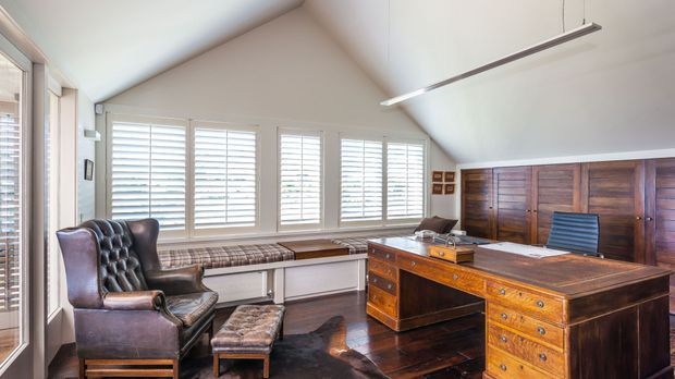 Bright, refined home office with a pitched ceiling, extensive plantation-shuttered windows, dark timber flooring, and substantial antique-style timber furniture. A large executive desk faces a tufted leather armchair and ottoman, with built-in timber cabinetry along the rear wall and a window seat beneath the glazing.