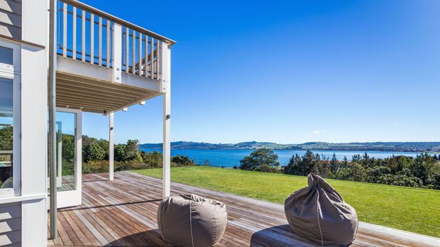 Exterior view of a contemporary multi-level home opening onto a broad timber deck and lawn, with an upper balcony featuring vertical timber or wood-toned balustrades. Two beanbag seats sit on the deck, overlooking a blue coastal or estuarine setting with distant low hills and islands.