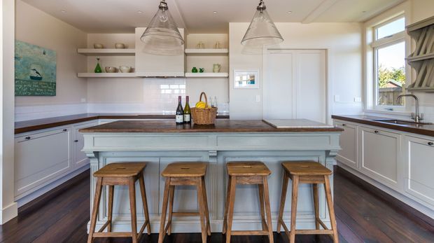 Bright, spacious traditional kitchen with a large pale blue-grey island, dark timber flooring, white shaker-style cabinetry, open shelving, and a dark stone benchtop. Four timber stools sit at the island, while pendant lights hang above. A window and glazed door bring in natural light, and decorative crockery and bottles add a lived-in feel.