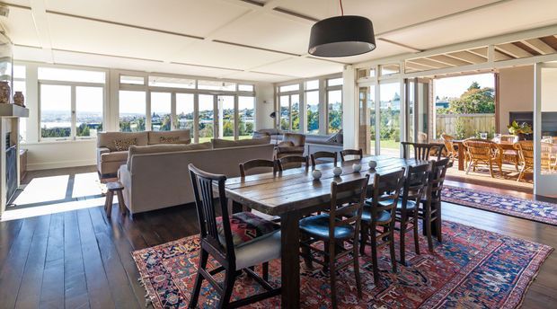 A bright, open-plan dining and living area with a large timber dining table and chairs arranged over layered patterned rugs. A sofa and armchairs occupy the adjoining lounge area, while extensive windows and glazed doors provide abundant daylight and views beyond. The room opens to a covered outdoor terrace with additional dining furniture. Dark timber flooring, neutral walls, exposed ceiling beams, and a large black pendant light define the interior.