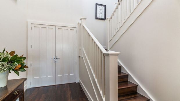 Bright traditional-style entry hall with a dark timber floor, white paneled double doors, and a white-painted staircase with turned balusters leading upward. A small framed artwork and a decorative console with flowers add detail to the otherwise light, refined space.