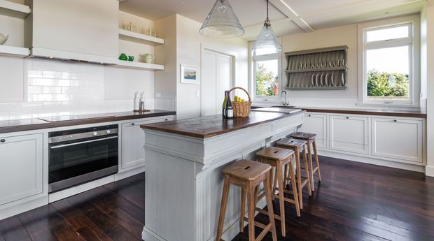 Bright traditional kitchen with a large central island, four timber stools, white panelled cabinetry, dark timber flooring, a dark stone benchtop, white subway-tile splashback, pendant lights, and a built-in open plate rack. The room has generous natural light from multiple windows and a clean, refined farmhouse-inspired character.
