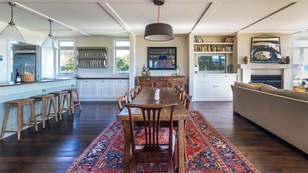Open-plan interior combining a kitchen, dining area, and living room. The space features dark timber flooring, pale built-in cabinetry, a long kitchen island with timber stools, a substantial timber dining table, a patterned Persian-style rug, pendant lighting, book shelving, and a fireplace with built-in joinery. Large windows provide abundant natural light.