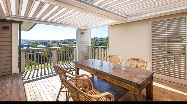 Covered outdoor dining terrace with a timber deck, large rustic timber dining table, wicker dining chairs, vertical timber balustrade, and a louvred pergola roof. The terrace overlooks a suburban coastal landscape and is connected to the home through timber-framed openings and louvred doors.