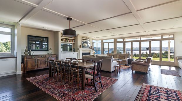 An open-plan dining and living area with dark timber flooring, a traditional wooden dining table and chairs set over patterned rugs, built-in shelving and cabinetry, and a large seating area facing expansive windows. The room has a coffered grid ceiling, pendant lighting, and bright views toward a landscaped exterior and distant water or sky.