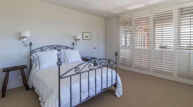 Bright, traditional-style bedroom with a white upholstered bed dressed in white linens and an ornate black metal bed frame. The room has beige carpet, pale walls, wall-mounted bedside lamps, a small wooden stool, and a wide bank of white plantation shutters covering the windows and glazed doors.