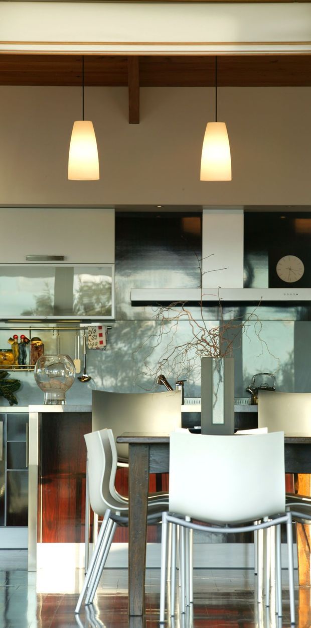 A contemporary open-plan kitchen and dining area with a high timber-lined ceiling, exposed timber beam, and two warm pendant lights. The kitchen features glossy dark cabinetry, reflective metallic splashback surfaces, pale upper cabinetry, a long stainless-steel rangehood, and a central island or dining counter. White modern bar stools and chairs surround a dark timber table, while sunlight and reflected greenery create patterned light across the surfaces.