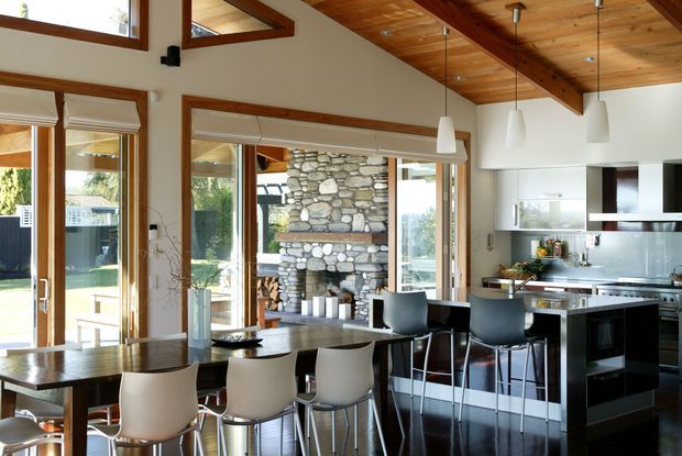 A bright, open-plan kitchen and dining area with large timber-framed windows and glass doors, a dark dining table, contemporary bar stools, white kitchen cabinetry, stainless-steel appliances, and a stone feature wall visible beyond the glazing. The room has a warm exposed timber ceiling with substantial beams and pendant lighting, creating a modern yet natural interior.