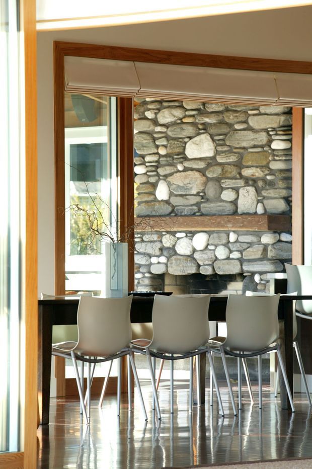 A bright contemporary dining area with a long dark dining table and streamlined white chairs, positioned in front of a large rustic stone fireplace. Full-height glazing and timber-framed openings bring in abundant natural light, while the polished dark floor reflects the furniture and stonework. A simple roller blind and sparse branch arrangement add understated detail.