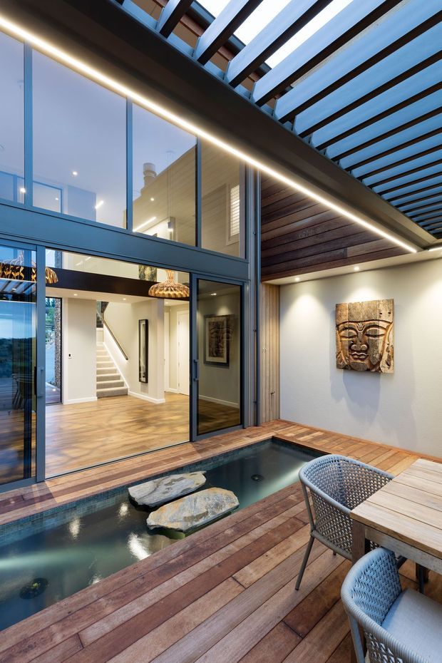A refined indoor-outdoor courtyard with a narrow plunge pool surrounded by timber decking. Full-height dark-framed glazing opens to a bright interior with timber flooring, a stairway and contemporary pendant lighting. The covered outdoor area features a timber-lined ceiling, exposed pergola structure, integrated linear lighting, outdoor dining furniture and a decorative wall carving.