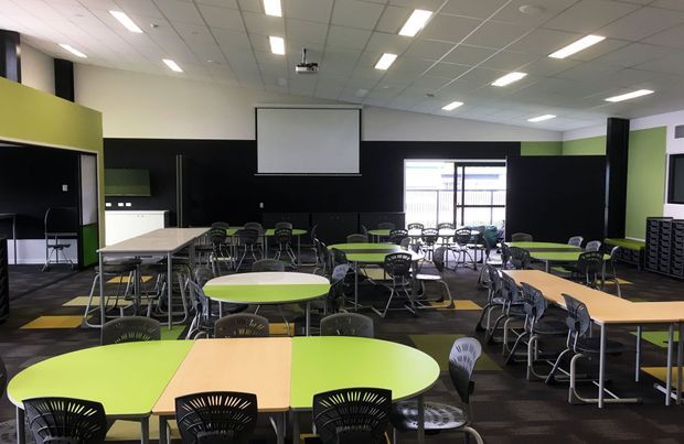 A large contemporary school or training classroom with numerous grouped tables and chairs, including bright lime-green and light timber tabletops. The room has black feature walls, lime-green accent walls, carpeted flooring, extensive recessed rectangular ceiling lighting, a ceiling-mounted projector, and a large projection screen. Windows and glazed doors at the rear provide natural light, while storage and presentation equipment line the walls.