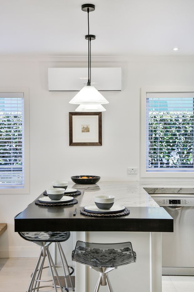 Bright contemporary kitchen with a compact marble-topped island or breakfast bar, dark waterfall-style edge, transparent bar stools, white cabinetry, stainless-steel appliances, plantation-shutter windows, and two white pendant lights. The island is set for casual dining with bowls and place settings.