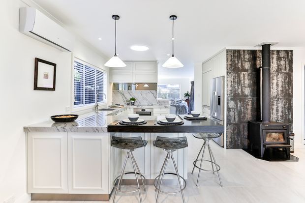 Bright open-plan kitchen with a white paneled island, dark stone benchtop, integrated appliances, pendant lights, and three bar stools. A dining and living area is visible beyond the kitchen, while a dramatic dark textured feature wall and freestanding wood-burning stove add contrast on the right. The room has white cabinetry, pale flooring, and abundant natural light.