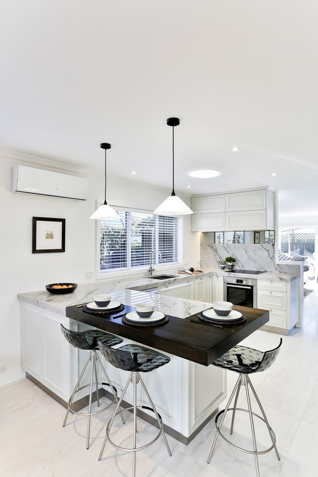 Bright contemporary white kitchen with a central marble-look island, integrated sink and cooktop, white shaker-style cabinetry, stainless steel appliances, and a dark timber breakfast bar with four transparent bar stools. Large windows with white blinds, pendant lights, recessed lighting, and a wall-mounted split-system air conditioner complete the space.