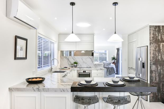 Bright, compact contemporary kitchen with white shaker-style cabinetry, a marble-look splashback and benchtop, a dark island with seating for three, stainless steel appliances, pendant lights, and a bedroom visible beyond the kitchen.