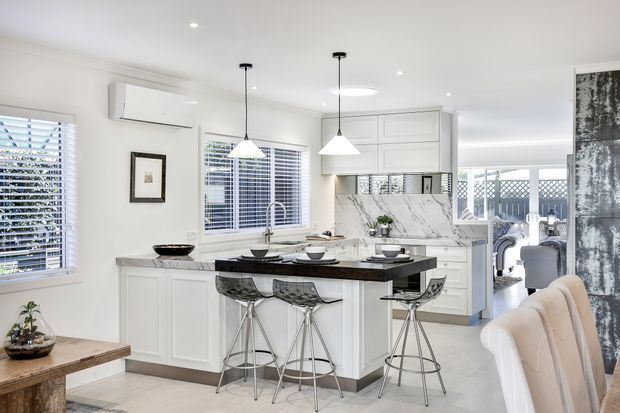 Bright, refined contemporary kitchen with a large central island, white paneled cabinetry, dark stone benchtop, marble-look splashback, pendant lighting, and stools. Plantation shutters, pale flooring, and an adjoining dining area create an airy open-plan setting.
