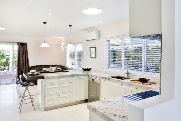Bright open-plan white kitchen with a central island, white shaker-style cabinetry, pale tiled flooring, marble-look benchtops, stainless-steel sink and dishwasher, and a dark cooktop. Pendant lights hang above the island, while a dining/living area and glazed doors are visible beyond. White plantation shutters and abundant daylight create a clean, airy interior.