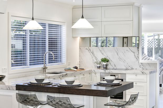 Bright white kitchen with shaker-style cabinetry, a large dark wood-toned island with seating, a pale marble-look backsplash and benchtops, stainless-steel fixtures, plantation shutters, and pendant lighting. The space opens toward a bright outdoor area through glazed doors.