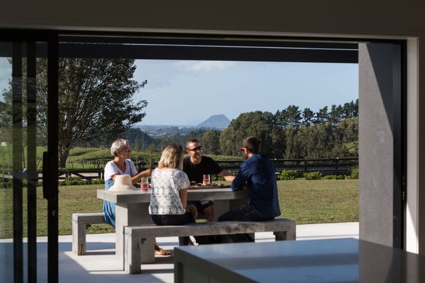 A contemporary indoor-outdoor dining area viewed through wide dark-framed glazing, with a concrete dining table and benches occupied by four people. Beyond the terrace is an open lawn, trees, distant hills and a rural landscape. A dark built-in counter or island is visible in the foreground.