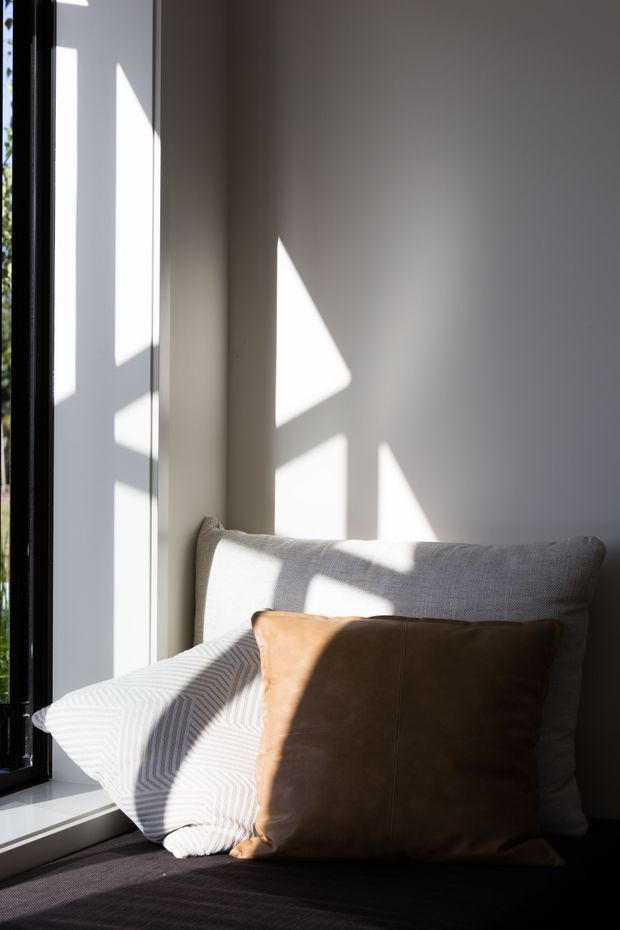 Close-up interior detail of a softly furnished window seat or bed corner, with neutral cushions and a warm brown accent pillow. Strong sunlight enters through the black-framed window, casting geometric shadows across the pale wall and textiles. The image has a minimalist, calm and intimate atmosphere.