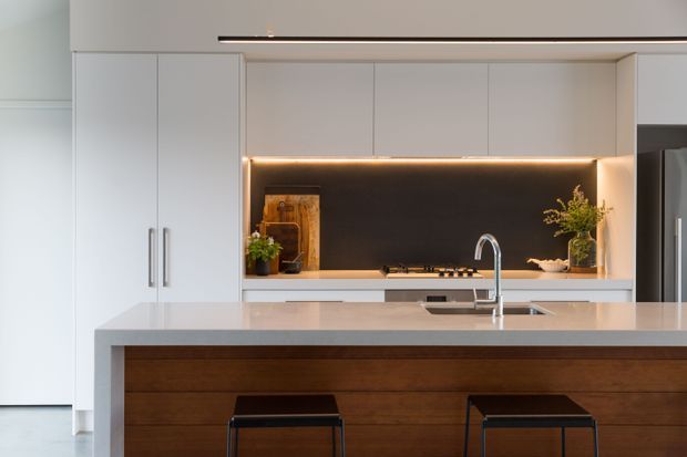 Contemporary minimalist kitchen with white floor-to-ceiling cabinetry, a large pale stone island benchtop, integrated sink and chrome tapware, and a warm dark timber island front. The rear cooking zone features a dark stone or glass splashback with concealed warm LED strip lighting and subtle decorative styling. Two black bar stools sit at the island.