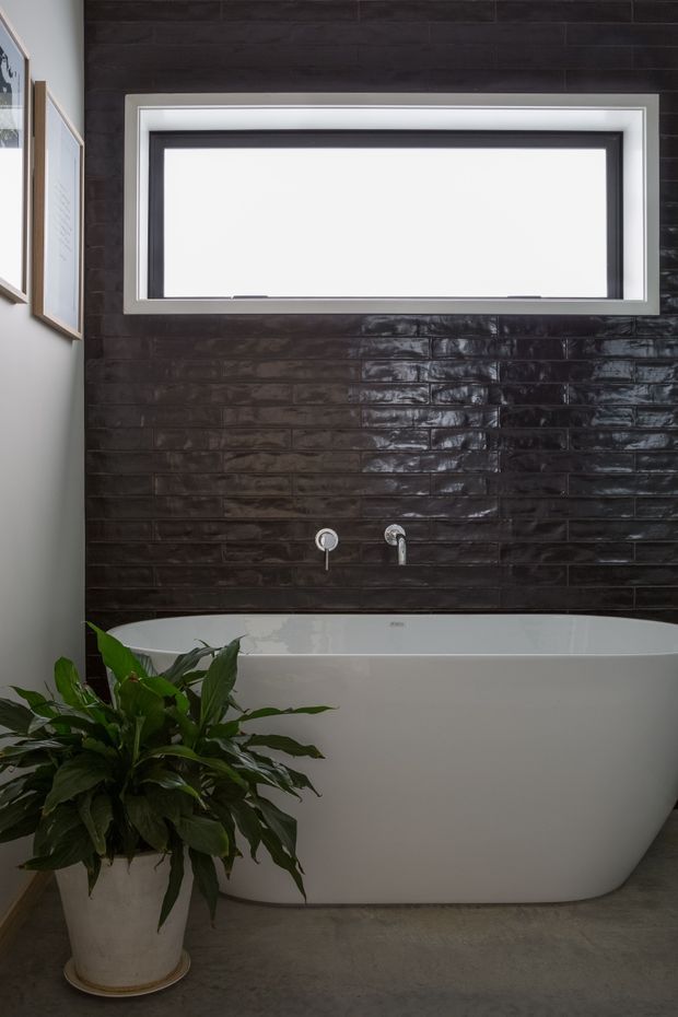 A modern bathroom featuring a freestanding white bathtub positioned against a dark glossy tiled feature wall. A wide horizontal window sits above the tub, with minimalist chrome wall-mounted fixtures. The room has a grey concrete-look floor, white walls, framed artwork at the side, and a large leafy potted plant in the foreground.