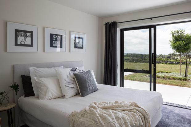A bright, softly styled bedroom with a neatly made upholstered bed, layered white and grey bedding, framed monochrome artwork, and dark curtains beside large black-framed sliding glass doors. The doors open to a paved terrace and green rural landscape beyond.