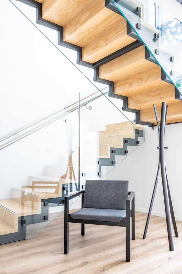 Bright contemporary interior featuring a sculptural floating staircase with warm timber treads, dark structural stringers, and clear glass balustrades with black fittings. A grey upholstered armchair and minimalist coat stand sit on pale timber flooring in the stair hall.