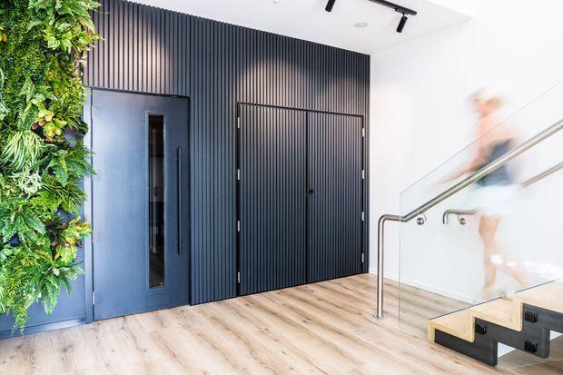 Bright contemporary interior entryway with light timber flooring, a dramatic black vertically ribbed feature wall and concealed double doors, a dark blue-grey entry door, a lush vertical garden wall, and a glass-and-metal stair balustrade leading to a lower level. A blurred person is moving on the stairs.