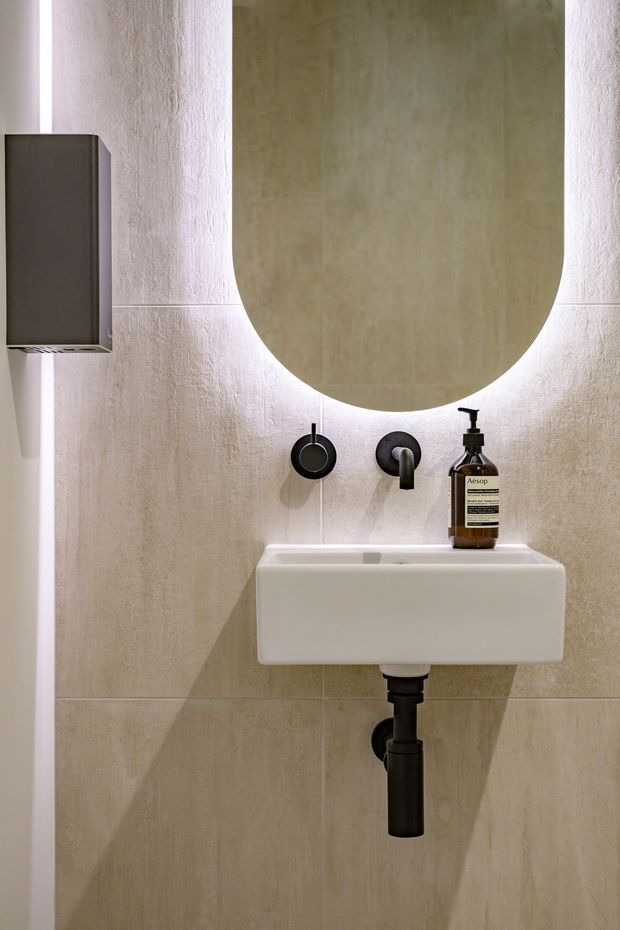A compact, minimalist bathroom featuring a wall-mounted white basin with exposed black plumbing, a black wall-mounted tap, a large rounded rectangular mirror with backlighting, pale textured stone-look wall tiles, and a dark wall-mounted storage unit. Warm concealed lighting creates a refined, contemporary atmosphere.